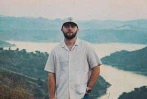 A man standing outdoors with a scenic lake and mountains in the background, during the California St.